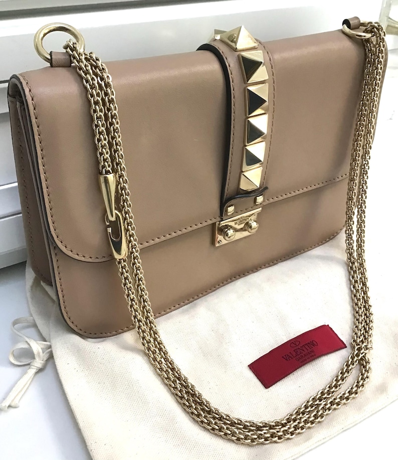 Op de afbeelding: Beige leren handtas met goudkleurige kettingriemen en piramidevormige studs. De klepsluiting heeft een gouden sluiting. Een rood label met witte tekst is zichtbaar.