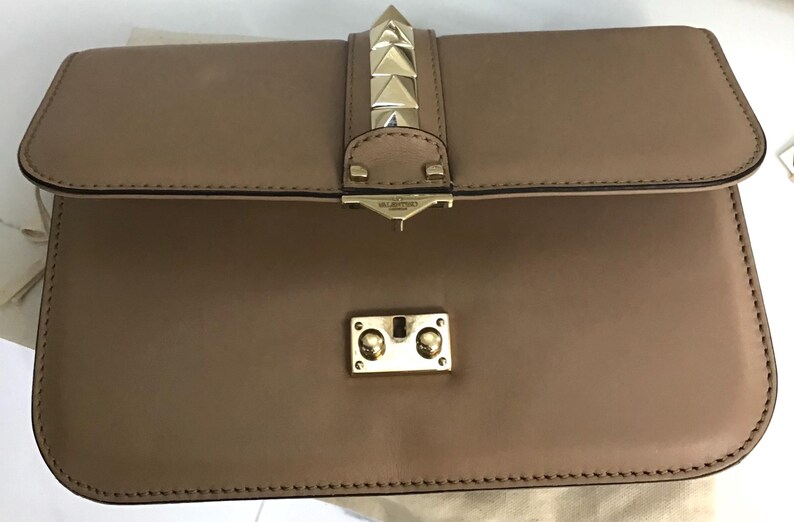 Op de afbeelding: Een bruinleren clutch met een klepsluiting. De clutch heeft goudkleurige piramidevormige studs aan de voorkant en een goudkleurige sluiting. De tas is rechthoekig en heeft een gestikte rand.