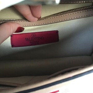 Op de afbeelding: Binnenaanzicht van een beige handtas met een rood Valentino Garavani label. Het label is rechthoekig met witte tekst en een logo. Een gouden rits en beige voering zijn ook zichtbaar.