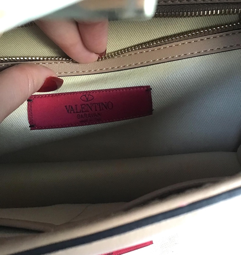Op de afbeelding: Binnenaanzicht van een beige handtas met een rood Valentino Garavani label. Het label is rechthoekig met witte tekst en een logo. Een gouden rits en beige voering zijn ook zichtbaar.