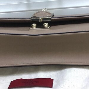 Op de afbeelding: Een taupekleurige handtas met een klepsluiting en goudkleurige hardware. De tas heeft een kettingriem en een rechthoekige vorm. De stiksels zijn zichtbaar langs de randen. Een rood label met witte tekst staat onderaan.