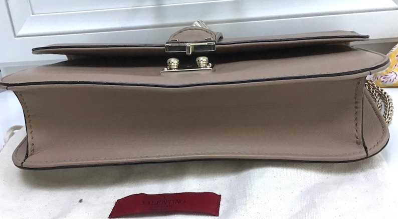 Op de afbeelding: Een taupekleurige handtas met een klepsluiting en goudkleurige hardware. De tas heeft een kettingriem en een rechthoekige vorm. De stiksels zijn zichtbaar langs de randen. Een rood label met witte tekst staat onderaan.