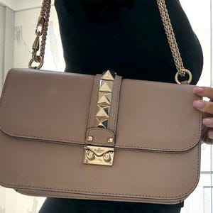 Op de afbeelding: Een beige rechthoekige handtas met een gouden kettingriem. De klepsluiting heeft gouden piramide studs en een gouden sluiting. De tas wordt vastgehouden door een persoon die een zwarte top draagt.