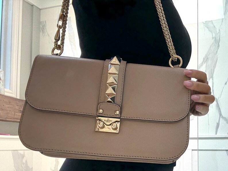 Op de afbeelding: Een beige rechthoekige handtas met een gouden kettingriem. De klepsluiting heeft gouden piramide studs en een gouden sluiting. De tas wordt vastgehouden door een persoon die een zwarte top draagt.