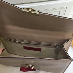 Op de afbeelding: Een beige leren clutch met een goudkleurige sluiting. De binnenkant heeft een ritsvak en een rood leren label. De tas is open en toont de binnenvoering en een klein rood label met de merknaam.