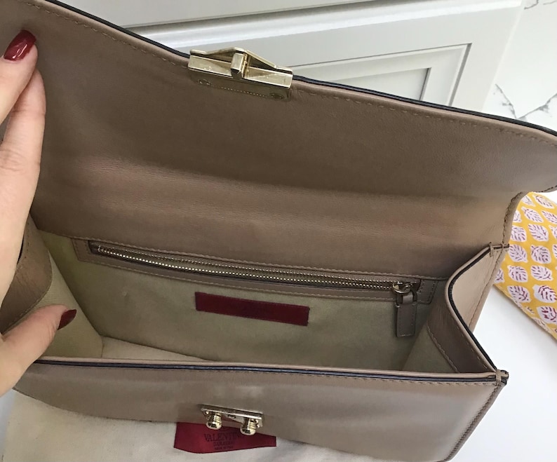 Op de afbeelding: Een beige leren clutch met een goudkleurige sluiting. De binnenkant heeft een ritsvak en een rood leren label. De tas is open en toont de binnenvoering en een klein rood label met de merknaam.
