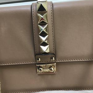 Op de afbeelding: Een bruinleren handtas met goudkleurige piramidevormige studs en een bijpassende sluiting. De tas heeft een kettingriem en is waarschijnlijk een schoudertas of crossbodytas.