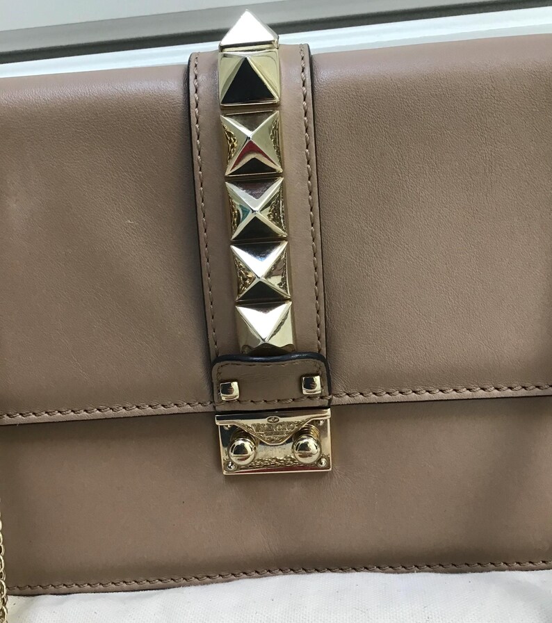 Op de afbeelding: Een bruinleren handtas met goudkleurige piramidevormige studs en een bijpassende sluiting. De tas heeft een kettingriem en is waarschijnlijk een schoudertas of crossbodytas.