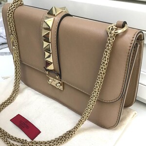 Op de afbeelding: Een beige leren schoudertas met een gouden ketting. De tas heeft gouden piramide studs op de flap en een gouden sluiting. Een rood label met de merknaam is zichtbaar.