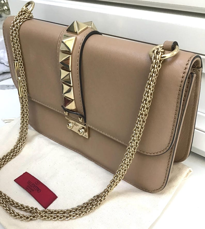 Op de afbeelding: Een beige leren schoudertas met een gouden ketting. De tas heeft gouden piramide studs op de flap en een gouden sluiting. Een rood label met de merknaam is zichtbaar.