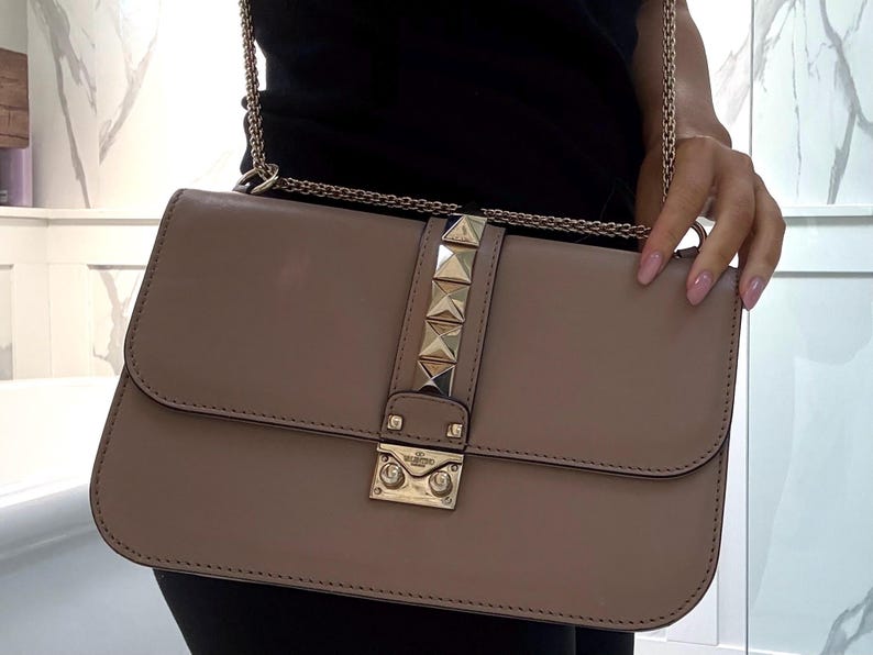Op de afbeelding: Een rechthoekige handtas in taupe met een gouden kettingriem. De tas heeft een gouden sluiting en een verticale gouden stud-accent. De tas wordt vastgehouden door een persoon die een zwarte top draagt.