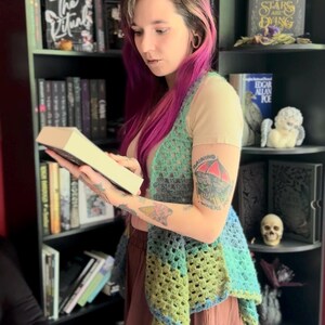 May include: A crochet vest in shades of green and blue, worn over a beige top and a brown skirt. The vest has a unique openwork design. A bookshelf filled with books is in the background.