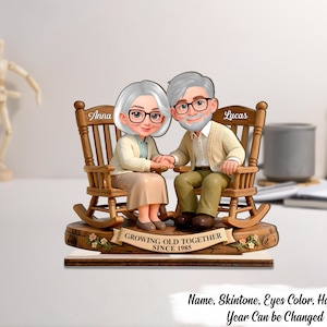 Puede incluir: Una figura de madera de una pareja de ancianos sentados en mecedoras, tomados de la mano. La mujer, llamada Anna, y el hombre, llamado Lucas, llevan gafas y sonríen. La base dice "GROWING OLD TOGETHER SINCE 1985".