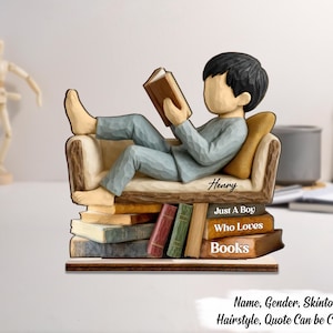May include: A wooden figurine of a person reading a book while reclining on a chair atop a stack of books. The figurine is wearing blue trousers and a long-sleeved shirt. The text on the figurine says "Henry", "Just A Boy Who Loves Books".