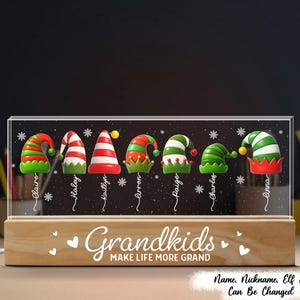 May include: A personalized night light featuring elf hats with names. The light has a wooden base and a clear acrylic panel with the words "Grandkids MAKE LIFE MORE GRAND". The text "Name, Nickname, Elf Hat Can Be Changed" is also visible.
