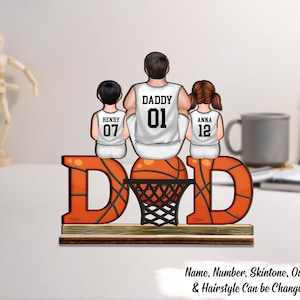 May include: A wooden basketball-themed family decoration. The word "DAD" is in orange, with a basketball and net in the center. Three figures in basketball jerseys are seated on the ball. The text "Name, Number, Skintone, Outfit & Hairstyle Can be Changed" is at the bottom.