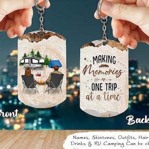May include: A keychain with a rustic design featuring a couple sitting by a campfire in front of a camper van. The back of the keychain has the text "Making Memories One Trip at a Time".