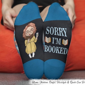 May include: A pair of black socks with blue toes. One sock features a cartoon of a person reading a book and holding a beverage. The other sock says "SORRY I'M BOOKED" with book graphics. The socks are on a person's feet.