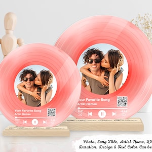Puede incluir: Dos marcos de fotos rosas con forma de disco de vinilo con una foto de una pareja. Los marcos se apoyan en bases de madera. El texto en el marco dice "Your Favorite Song, Artist Names" e incluye un código QR. También es visible el texto "Photo, Song Title, Artist Name, QR Code, Duration, Design & Text Color Can be Changed".