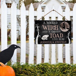 Puede incluir: Un letrero de Halloween en blanco y negro con una bruja y un sapo sobre un fondo negro. El letrero dice "A Wicked Witch and a Grumpy Toad Live Here Jennifer & Jonathan".