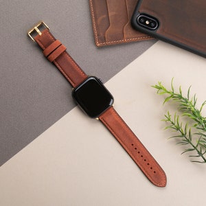 May include: A brown leather watch band with a gold buckle. The band is shown on a white surface with a brown leather wallet and a brown leather phone case in the background.