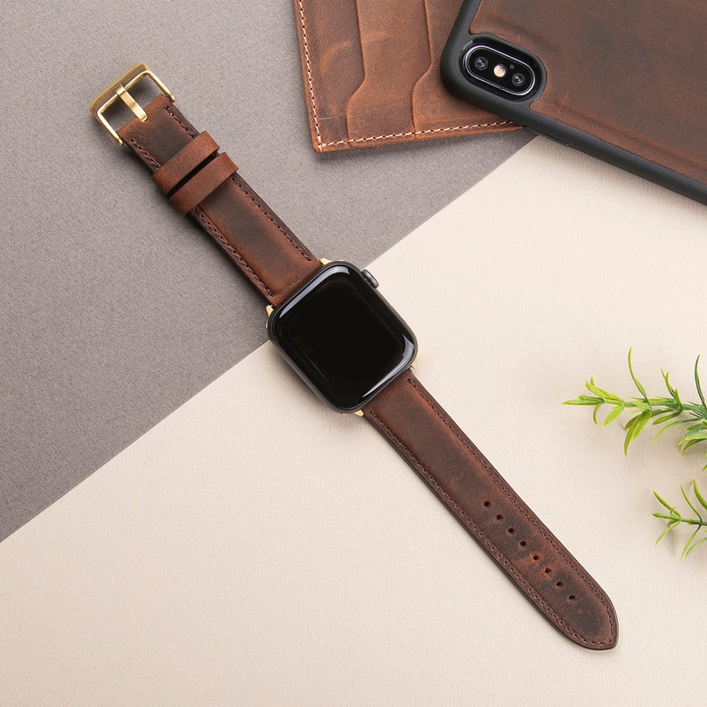 May include: A brown leather watch band with a gold buckle and a black Apple Watch face.