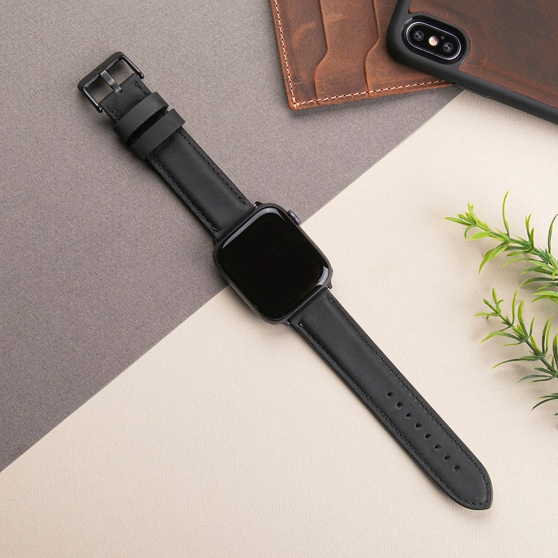 May include: A black leather watch band with a black watch face. The band has a buckle closure and is shown on a light background.