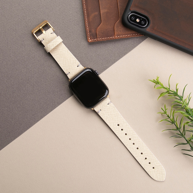 May include: A white leather watch band with a gold buckle and a black Apple Watch face.