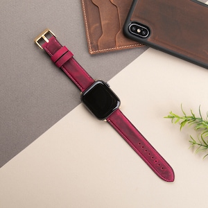 May include: A burgundy leather watch band with a gold buckle and a black Apple Watch face.