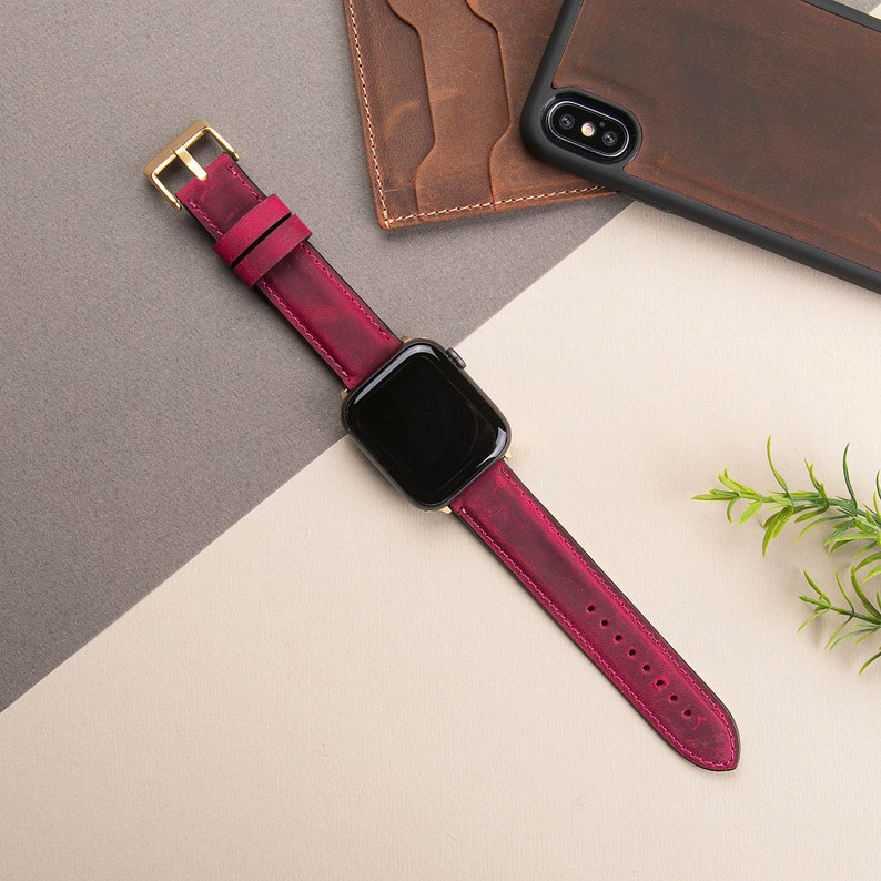 May include: A burgundy leather watch band with a gold buckle and a black Apple Watch face.