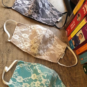 May include: Three face masks with white lace patterns on different colored fabric backgrounds. The masks are gray, beige, and teal.