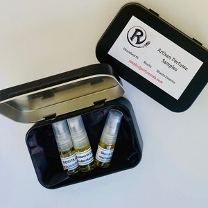 May include: Black metal tin containing three small perfume sample bottles. The bottles are filled with a golden liquid. The tin's lid displays a label with the text "Artisan Perfume Samples" and the brand name "Rosebud Perfume Lab".