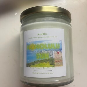 May include: AmmBer candle in a glass jar with a gold lid. The label reads "HONOLULU SUN" with a scenic image. The text "Made with soy wax and essential oils" and "Hand Poured in Small Batches" are also visible.