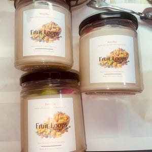 May include: Four glass jars of scented candles with black lids. Each jar has a white label with the text "Fruit Loops" and a graphic of a bowl of colorful cereal. The candles are hand-poured and made with soy wax.