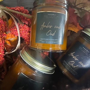 May include: Three amber glass candle jars with black labels. The labels have gold lettering that reads "AmmBer" and "Amber and Oud". The candles are hand-poured in small batches and made with love in Maryland.