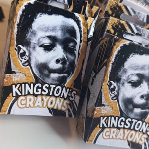 May include: Multiple rectangular cardboard boxes labeled Kingston's Crayons, each featuring a black and white portrait of a young child on the front. The packaging design incorporates gold and black abstract paint splatter accents on the edges.