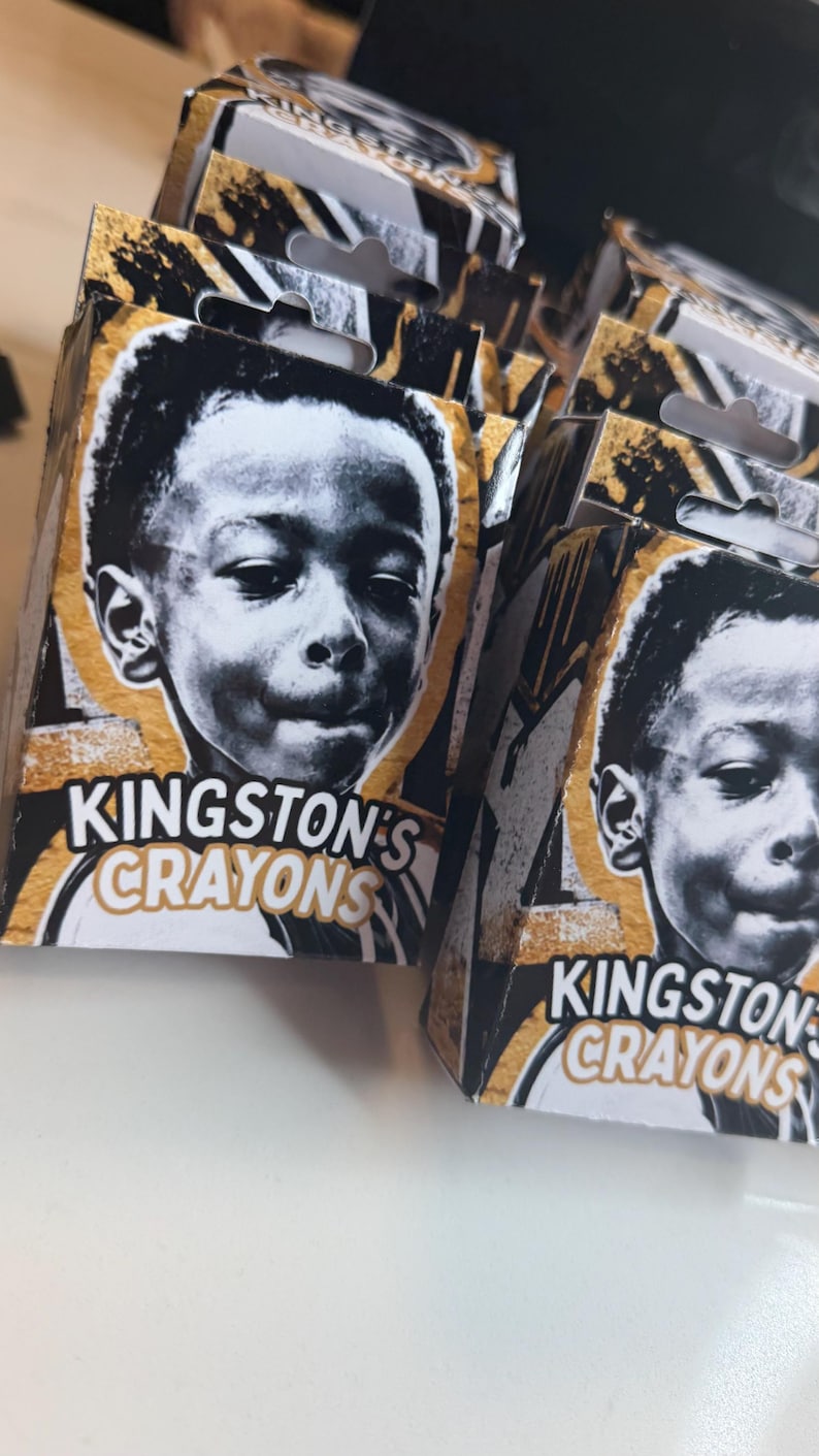 May include: Multiple rectangular cardboard boxes labeled Kingston's Crayons, each featuring a black and white portrait of a young child on the front. The packaging design incorporates gold and black abstract paint splatter accents on the edges.