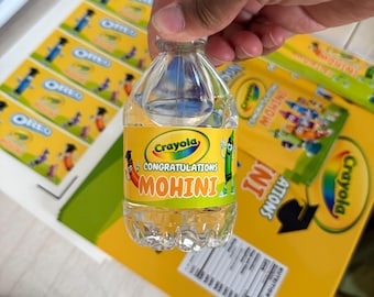 May include: A small, clear plastic water bottle with a yellow label featuring the Crayola logo and the text "Congratulations MOHINI." The bottle is held by a hand, with Oreo labels and a yellow Crayola box in the background.