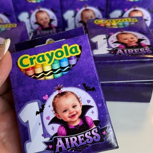 May include: A purple Crayola crayon box featuring a graphic of a baby dressed as a vampire with the text 1 AIRESS. The box displays a cutout window showing colorful crayons inside, with a large number one and small bats and hearts surrounding the baby.