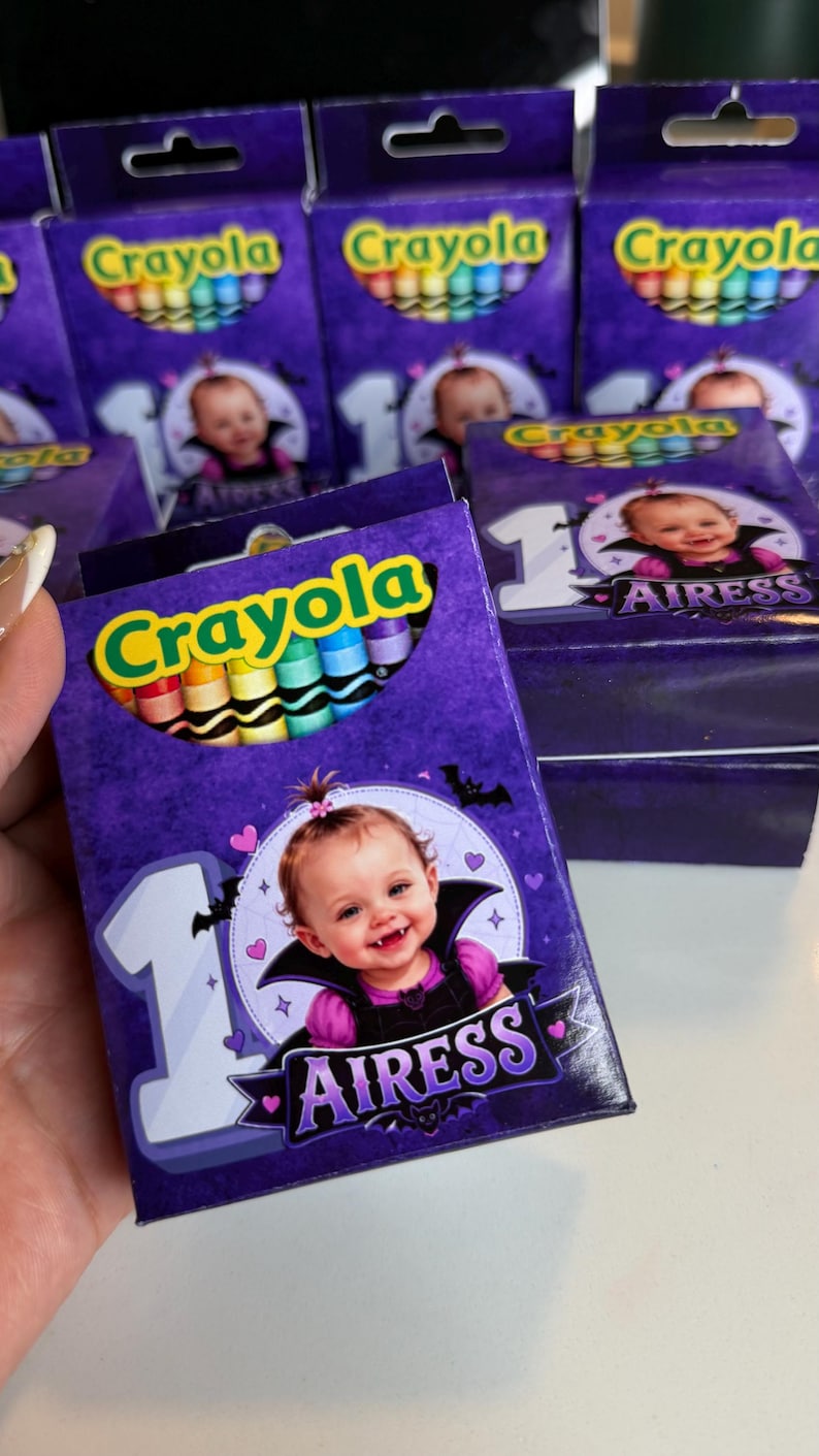 May include: A purple Crayola crayon box featuring a graphic of a baby dressed as a vampire with the text 1 AIRESS. The box displays a cutout window showing colorful crayons inside, with a large number one and small bats and hearts surrounding the baby.