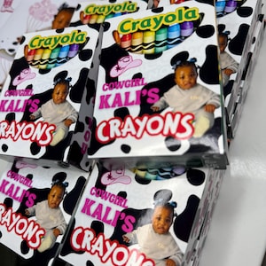 May include: Multiple rectangular boxes of Crayola crayons featuring a black and white cow print pattern. Each box displays a photograph of a young child, a pink cowgirl hat icon, and the text Cowgirl Kali's Crayons in bold pink and red lettering.