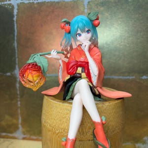 Può includere: Una statuina di un personaggio con capelli turchesi e accenti rossi, che indossa un kimono e tiene in mano una lanterna decorativa. La figura è seduta su un contenitore in stile bambù, con sandali rossi. Lo sfondo ha un motivo dorato.