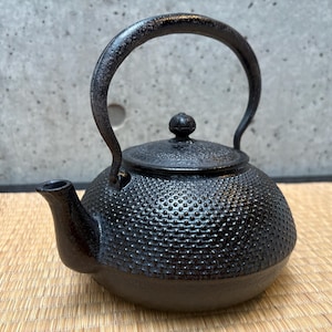 Puede incluir: Tetera de hierro fundido negro con una superficie texturizada y con hoyuelos. La tetera tiene un asa curva y un pequeño pico. La tapa está rematada con un pequeño pomo. La tetera está diseñada para preparar té.