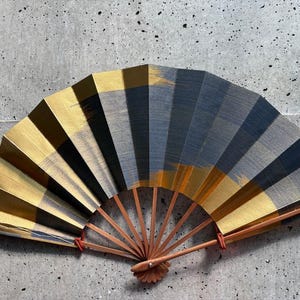 May include: A folding hand fan featuring a pleated surface with alternating panels of metallic gold and dark charcoal grey. The fan is constructed with thin, light brown wooden ribs that radiate from a central pivot point at the base.