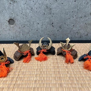 Puede incluir: Cinco cascos de samurái en miniatura con detalles intrincados. Los cascos presentan acentos negros, marrones y dorados, con borlas naranjas vibrantes. Los cascos se exhiben sobre una estera tejida, mostrando la artesanía japonesa.