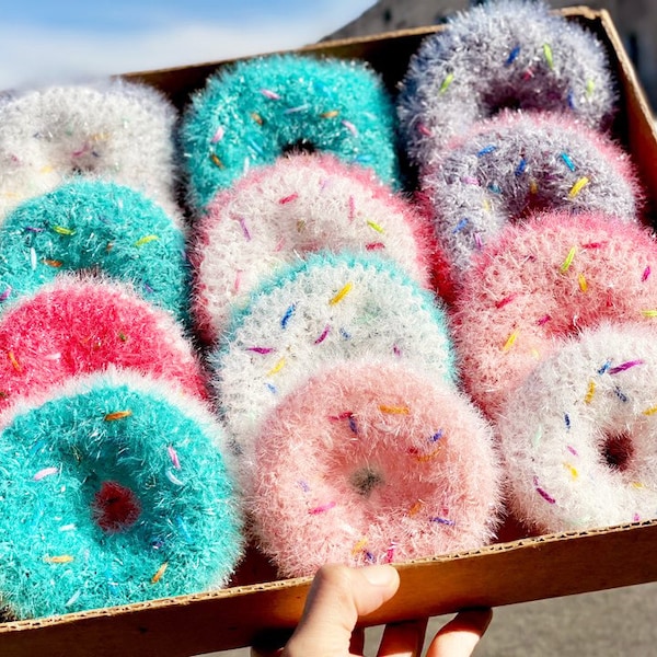 May include: A cardboard box filled with a dozen handmade donut-shaped scrubbies. The scrubbies are in various colors, including pink, blue, white, and gray, with colorful sprinkles. The scrubbies are fluffy and textured.