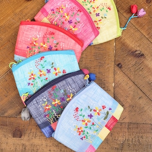 May include: A collection of colorful, embroidered coin purses. Each purse features a different color, including pink, blue, and yellow, with a floral design. The purses have a zipper closure and a small tassel or pom-pom attached. They are arranged on a wooden surface.