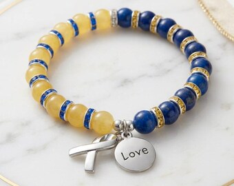 May include: A beaded bracelet with yellow and blue stones. The bracelet has a silver charm with the word "Love" and a ribbon. A gold ring and a white dish with a gold rim are in the background.