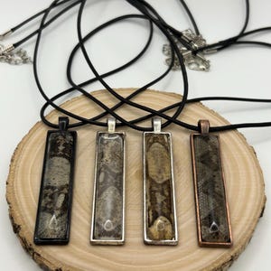May include: Four rectangular pendant necklaces with snake skin patterns. The pendants are set in black, silver, and copper-colored frames, each suspended from a black cord. The necklaces are displayed on a wooden slice.
