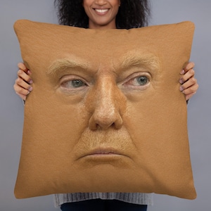 May include: A square decorative pillow featuring a realistic image of a face. The face has light skin, blonde hair, and a serious expression. The pillow is a light tan color.